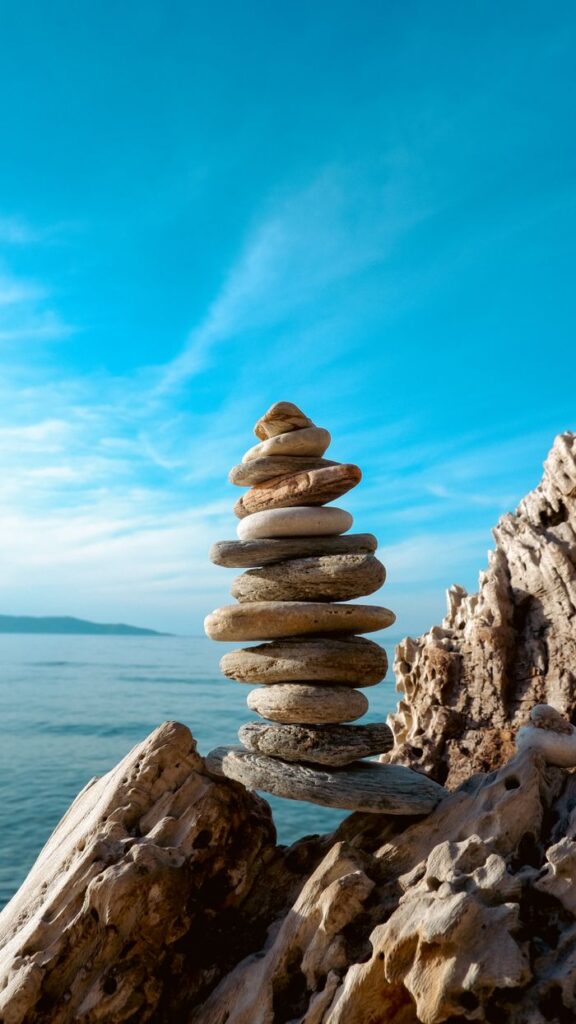 A stack of balanced stones on a rocky seashore with a clear blue sky and sea in the background.