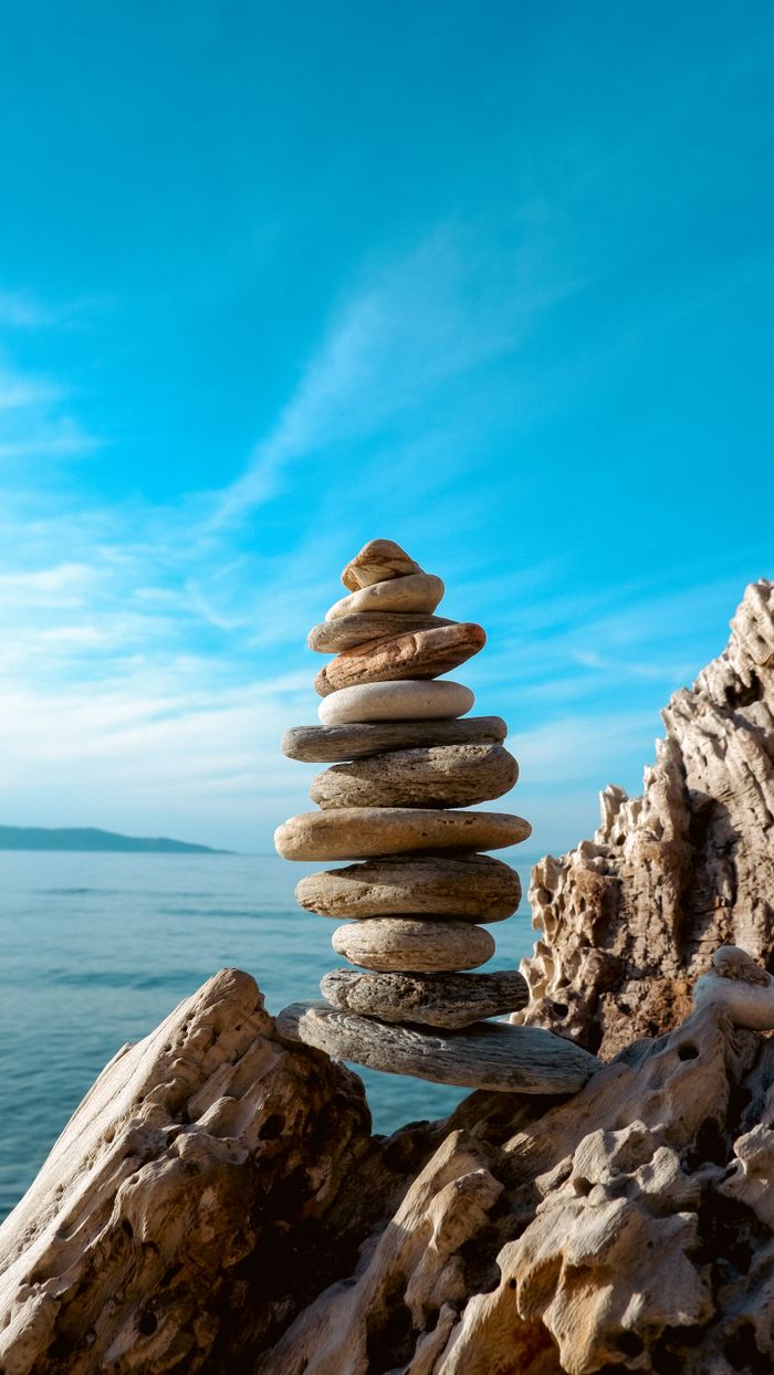 The Art of Drawing Readers In: Your attractive post title goes here A stack of balanced stones on a rocky seashore with a clear blue sky and sea in the background.